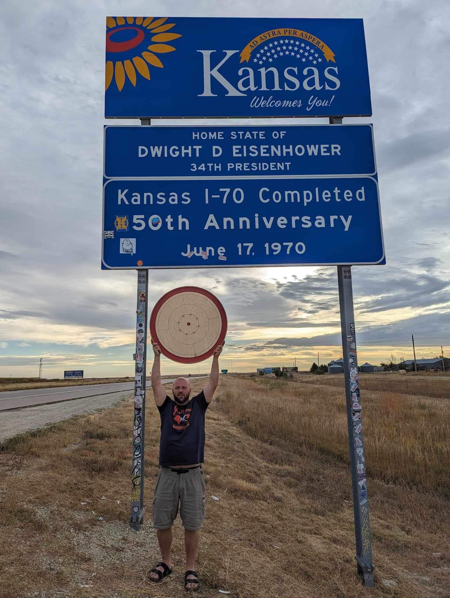 Jeremy Tracey poses with crokinole board before Kansas sign on highway